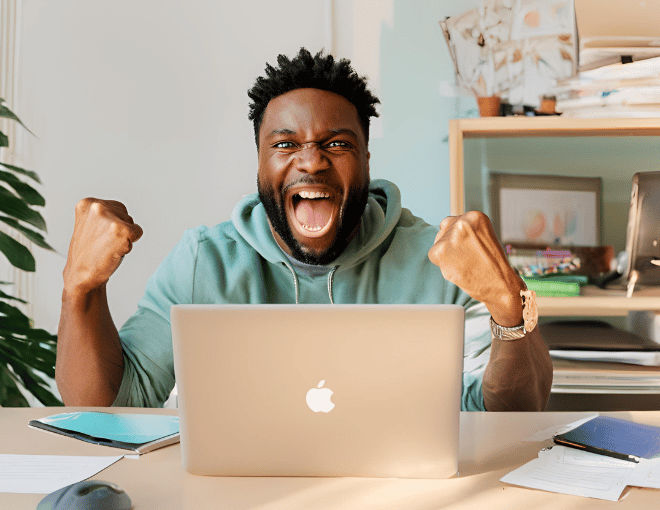 A man jubilantly raises his fists, eyes gleaming with joy, while sitting at a desk, a glowing laptop screen displaying his success.