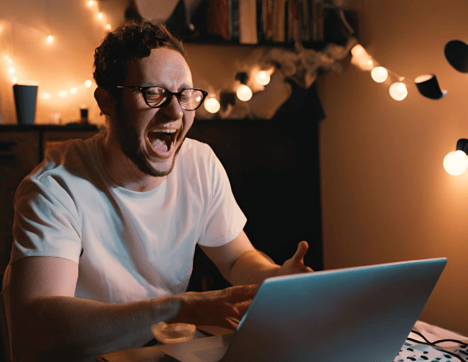 A white man cheers in his dimly lit room, laptop glowing, as he wins big in his room.