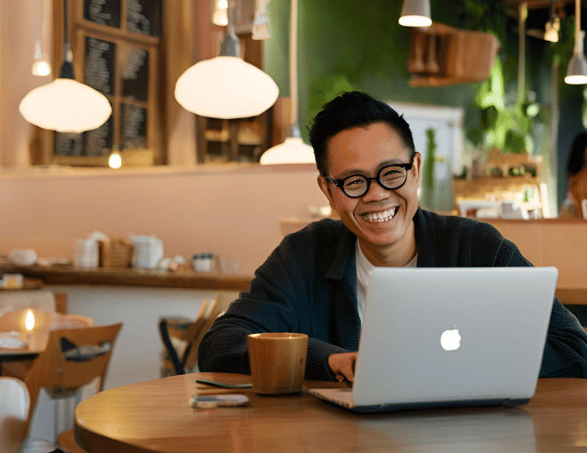 In a cozy café, an Indonesian man grins at his laptop, the light from the screen reflecting off his glasses.